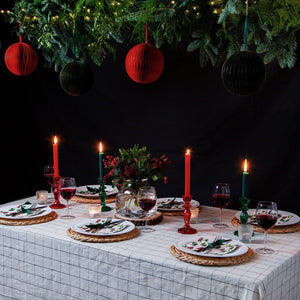Red and green glass candle holders on a Christmas dinner table with holly-themed plates, wine glasses, and hanging paper baubles.
