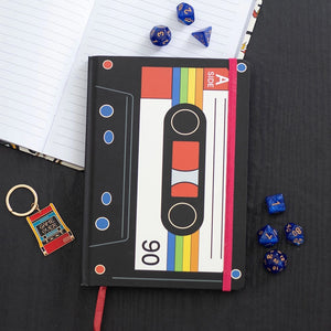 Closed notebook on desk with dice and keyring – Retro cassette tape A5 notebook with bold 80s design beside dice and accessories.