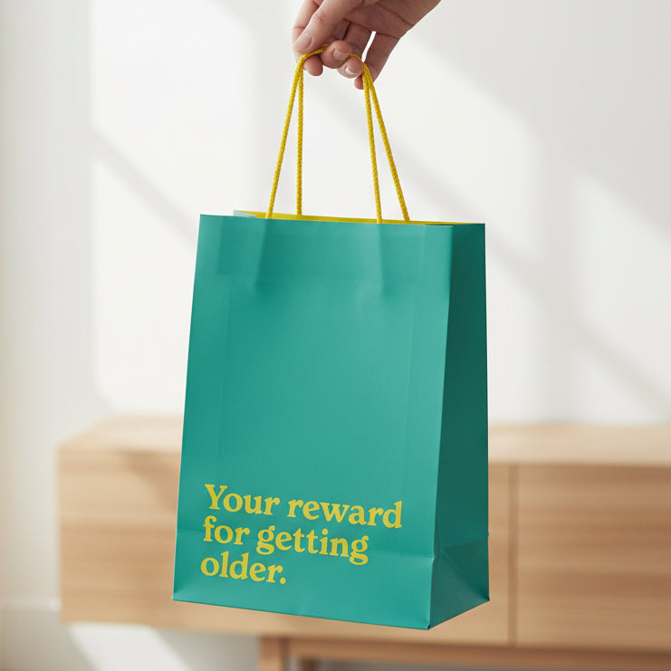 Teal slogan gift bag held in hand showing gold text and braided handles.