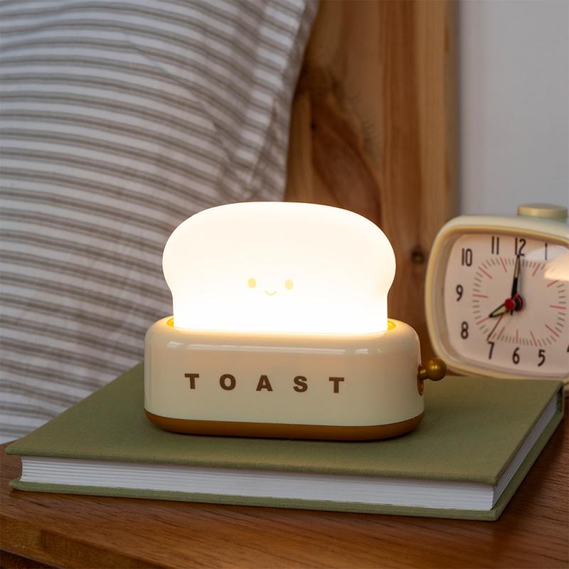 Close-up of Kawaii toaster night light glowing on a bedside table with books and alarm clock – USB rechargeable dimmable lamp.