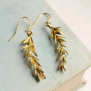 A pair of gold-toned rosemary leaf earrings resting on a pale blue book cover, showcasing their intricate detailing and elegant design.