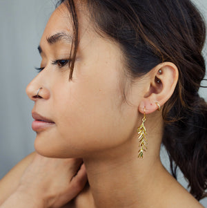 A woman wearing the gold rosemary leaf earrings, highlighting their graceful length and how they dangle just below the jawline.