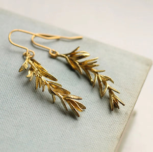 Close-up of the gold rosemary leaf earrings on a textured fabric background, displaying the delicate leaf pattern and craftsmanship.