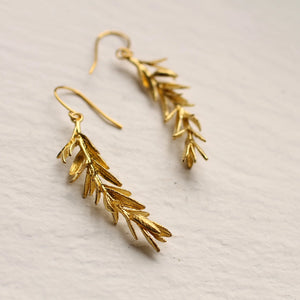 A pair of gold rosemary leaf earrings lying on a white surface, emphasizing their organic shape and shimmering brass finish.