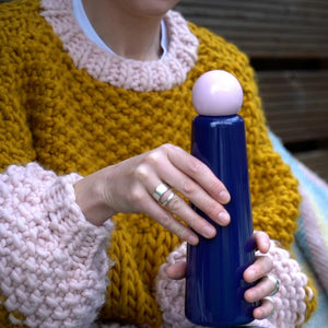 Person holding Skittle Bottle Indigo & Pink 750ml, stylish navy reusable water bottle with pink rounded lid, outdoor lifestyle setting.