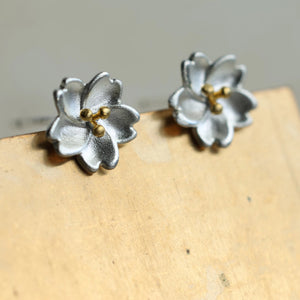Sterling silver cherry blossom stud earrings with gold centre details displayed on a wooden surface – delicate floral design jewellery