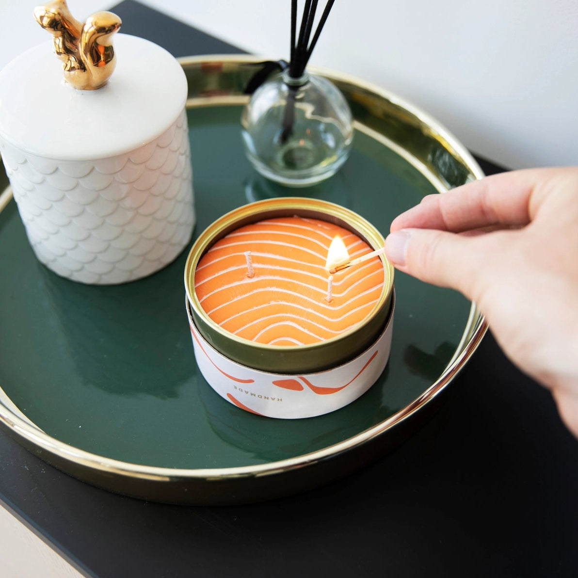 Hand lighting the Orange Salmon tin candle on a dark green tray with modern home décor in the background.
