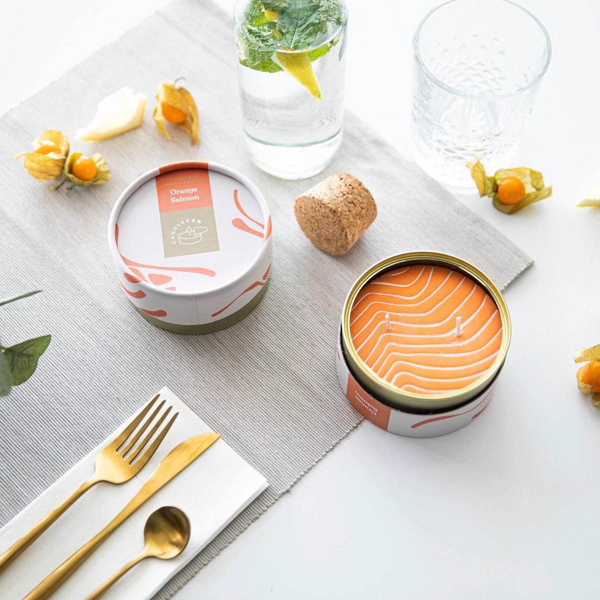 Flat-lay of Orange Salmon CandleCan with lid and open tin surrounded by fruit slices and glassware on a grey placemat.