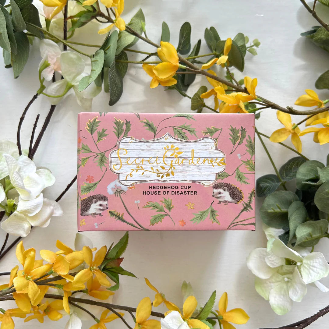 Secret Garden Hedgehog Cup packaging box surrounded by yellow flowers.
