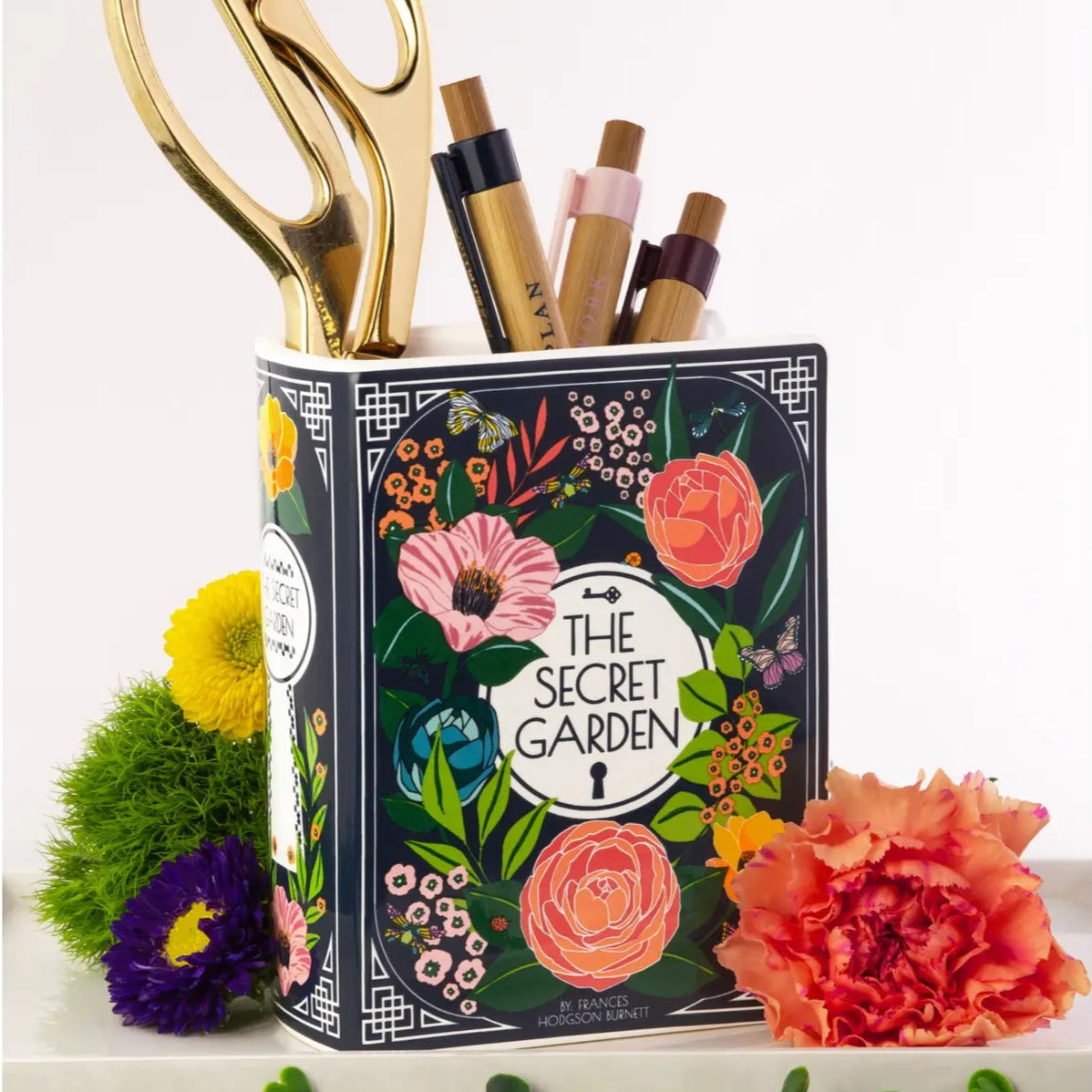 Secret Garden book vase used as a pen and scissor holder on a desk with flowers and props