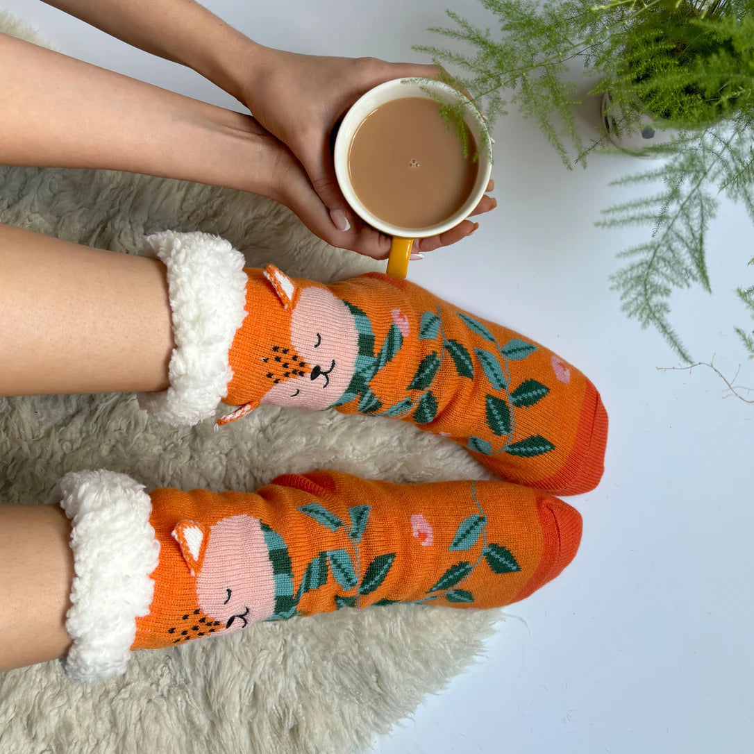 Person wearing orange Secret Garden Fox Slipper Socks while holding a cup of tea, with a fluffy rug and green fern in the background. Cosy autumnal setting.