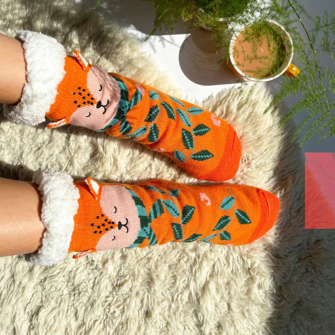 Brightly lit view of orange fleece-lined fox slipper socks with leafy forest pattern, worn by a person relaxing with a cup of tea on a cream rug.