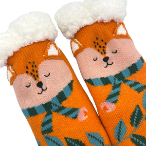 Close-up of fox faces on orange slipper socks with 3D ears and green stripy scarves, highlighting detailed knit and fleece lining.