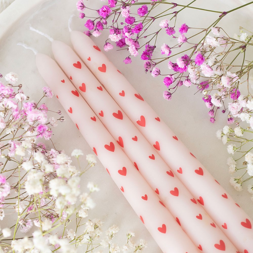 Four pink taper candles with red love hearts surrounded by pink and white gypsophila flowers, styled flat lay for romantic Valentine’s Day décor.