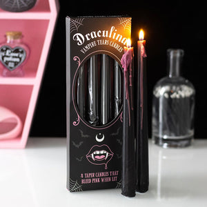 Set of pink-drip vampire taper candles burning next to gothic-themed packaging on a white surface with Halloween decor in background.
