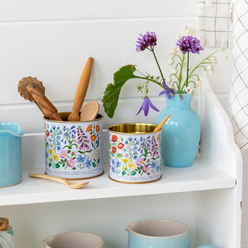 Set of two wildflower tins on white shelf with wooden utensils and flowers.
