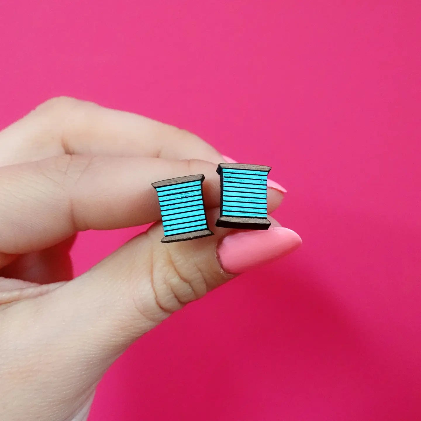 Front view of turquoise bobbin thread stud earrings held between fingers against a pink background – handcrafted wooden earrings for sewing lovers.