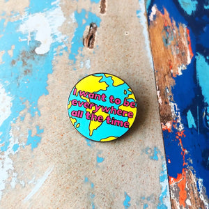 Silly Loaf Travel Enamel Pin featuring a colourful globe design with bold pink text reading ‘I want to be everywhere all the time’, placed on a rustic blue and wooden background.