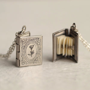 A silver book locket necklace displayed open and closed, revealing its hidden pages inside.