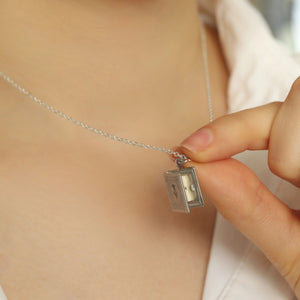 A hand holding the silver book locket, partially open, revealing its miniature pages.