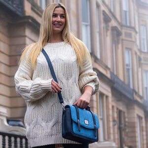 Woman wearing sky blue Harris Tweed messenger bag crossbody with navy strap and cream jumper in city backdrop.