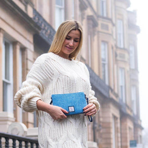 Woman holding sky blue Harris Tweed zip bag in urban setting, styled as a clutch with chunky white knit jumper.