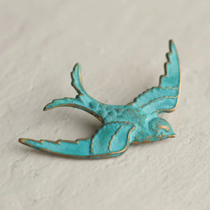 A small verdigris swallow brooch with a turquoise and gold patina, lying on a textured white surface.