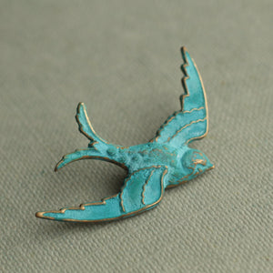 A close-up of the verdigris swallow brooch on a muted green fabric background, showcasing its intricate gold detailing.