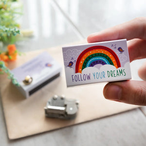 Hand holding a Follow Your Dreams matchbox gift with a rainbow design.