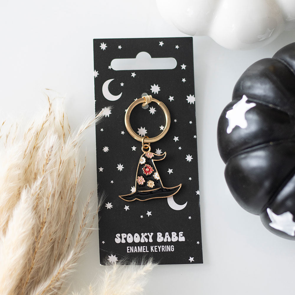 Spooky Babe enamel keyring on a black card with decorative elements, surrounded by pampas grass and a black pumpkin.