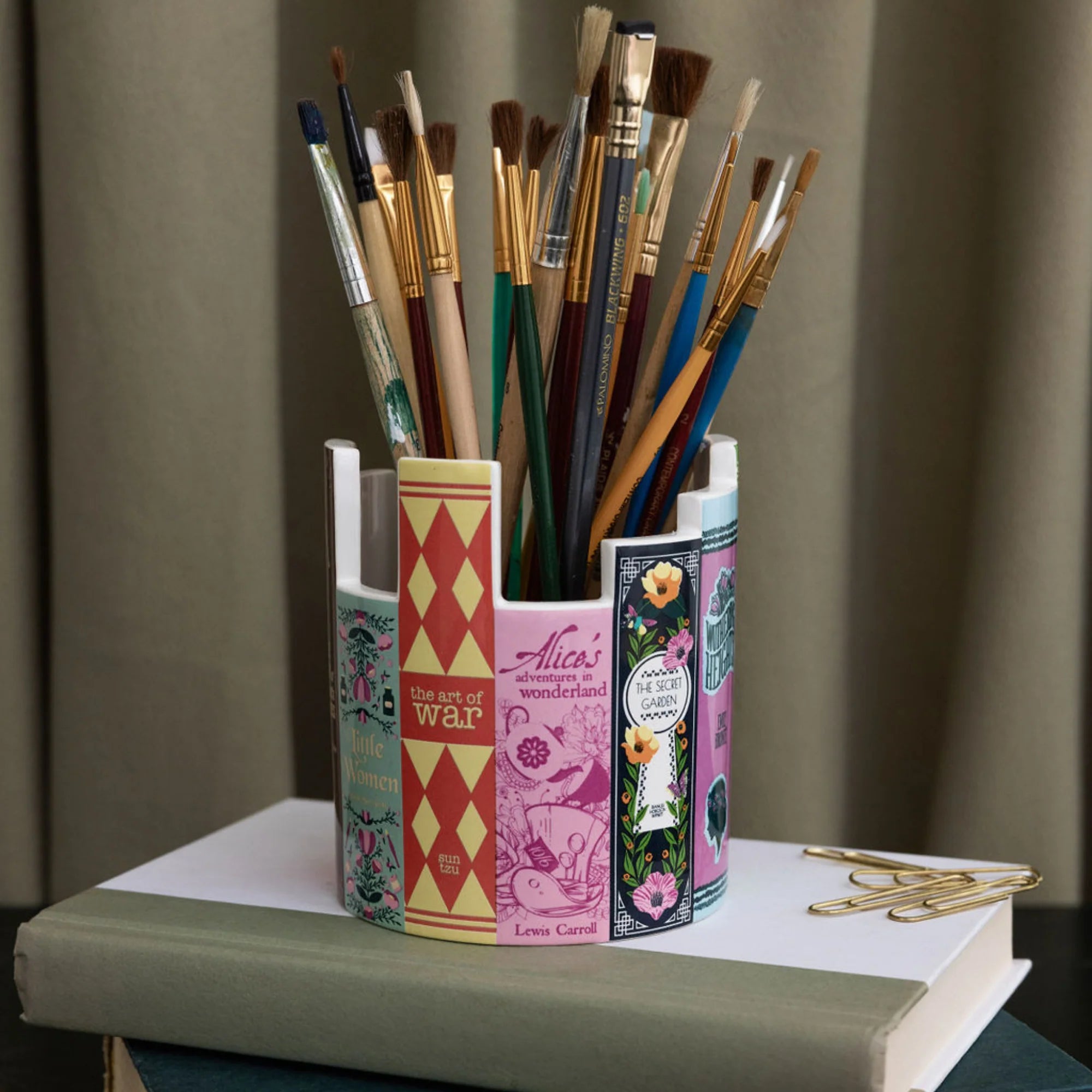 Ceramic literary book pot used as a paintbrush holder on a desk, surrounded by books, gold paperclips, and soft beige curtains.