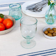 Blue stemmed bubble glass on a laid table with oranges, olives, and cutlery in the background.