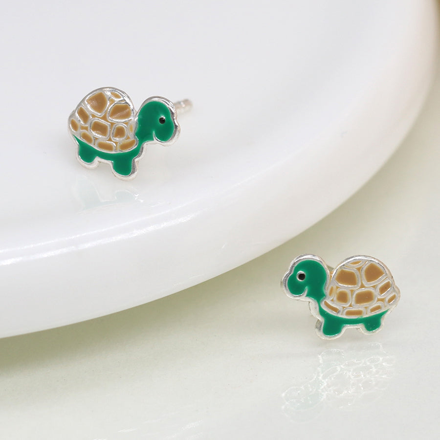 Close-up of sterling silver enamel turtle earrings displayed near a white ceramic dish, showcasing their tiny size and charming design.