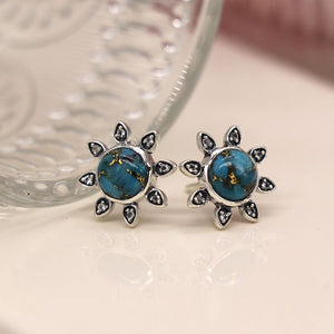 Close-up of a pair of sterling silver sunflower stud earrings with a turquoise and copper Mojave stone in the center and white topaz inset petals, resting on a wooden table top.