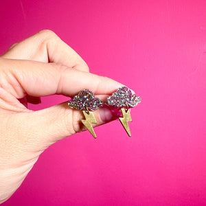 Hand holding a pair of sparkly storm cloud earrings with gold lightning bolts against a vibrant pink background.