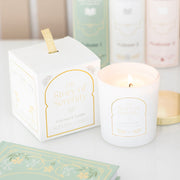 The Story of Serenity Coconut & Vanilla Candle lit in frosted white jar with gold lid, shown beside elegant illustrated gift box.