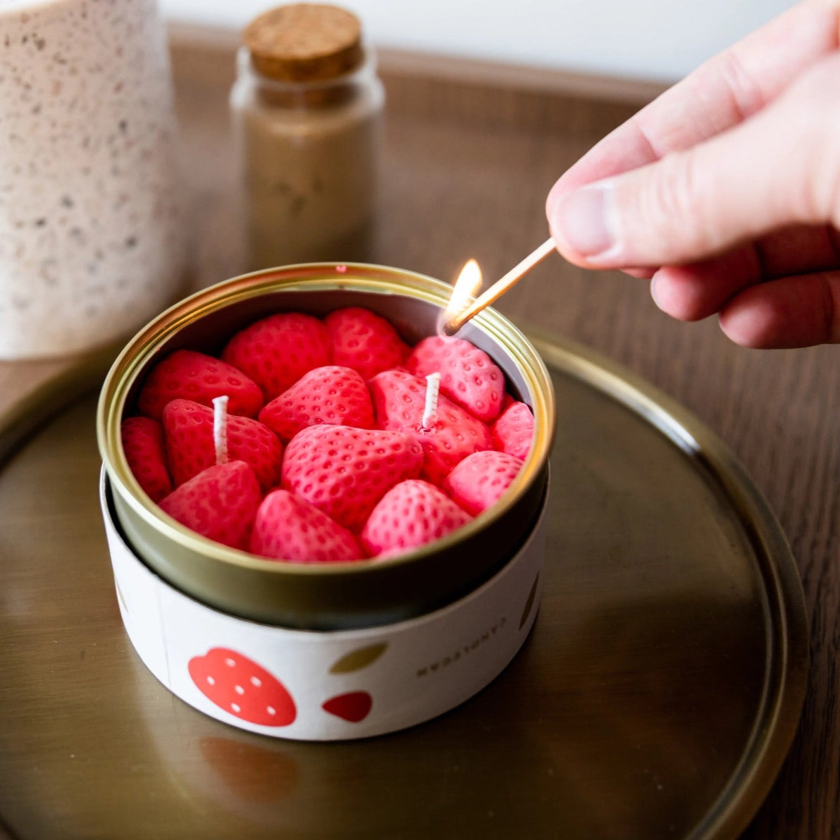 Lighting the Strawberry CandleCan with a match, displaying detailed strawberry-shaped wax pieces inside the round tin on a table.