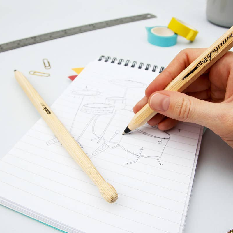 Person sketching a drum kit in a notebook using a realistic drumstick pen with black ink on a tidy desk.