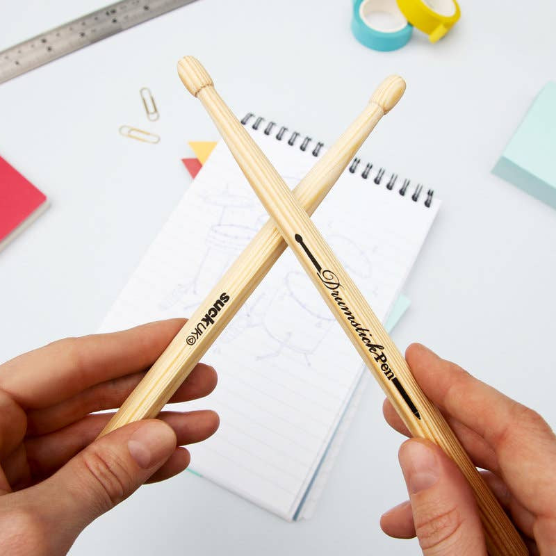 Two hands holding crossed Suck UK drumstick pens above a notepad, highlighting the pen’s natural wood finish and playful design.