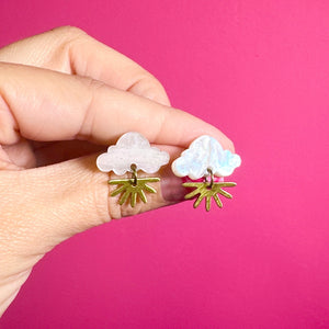 Close-up of hand holding a pair of cloud-shaped stud earrings with glittery white acrylic tops and dangling brass sunshine rays, set against a hot pink background.