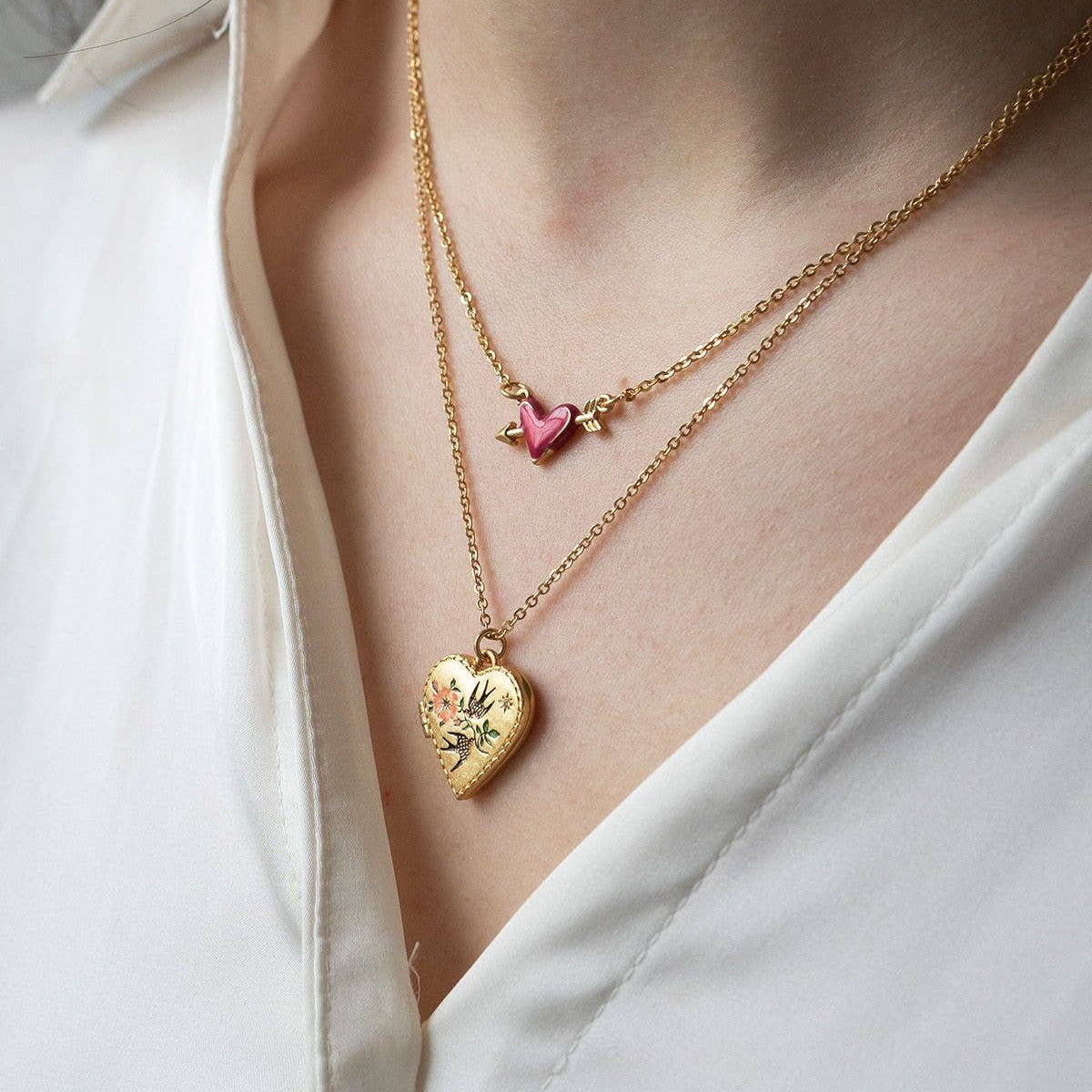Woman wearing layered gold necklaces, featuring the Fable England Swallow Heart Locket and a small pink heart charm necklace.