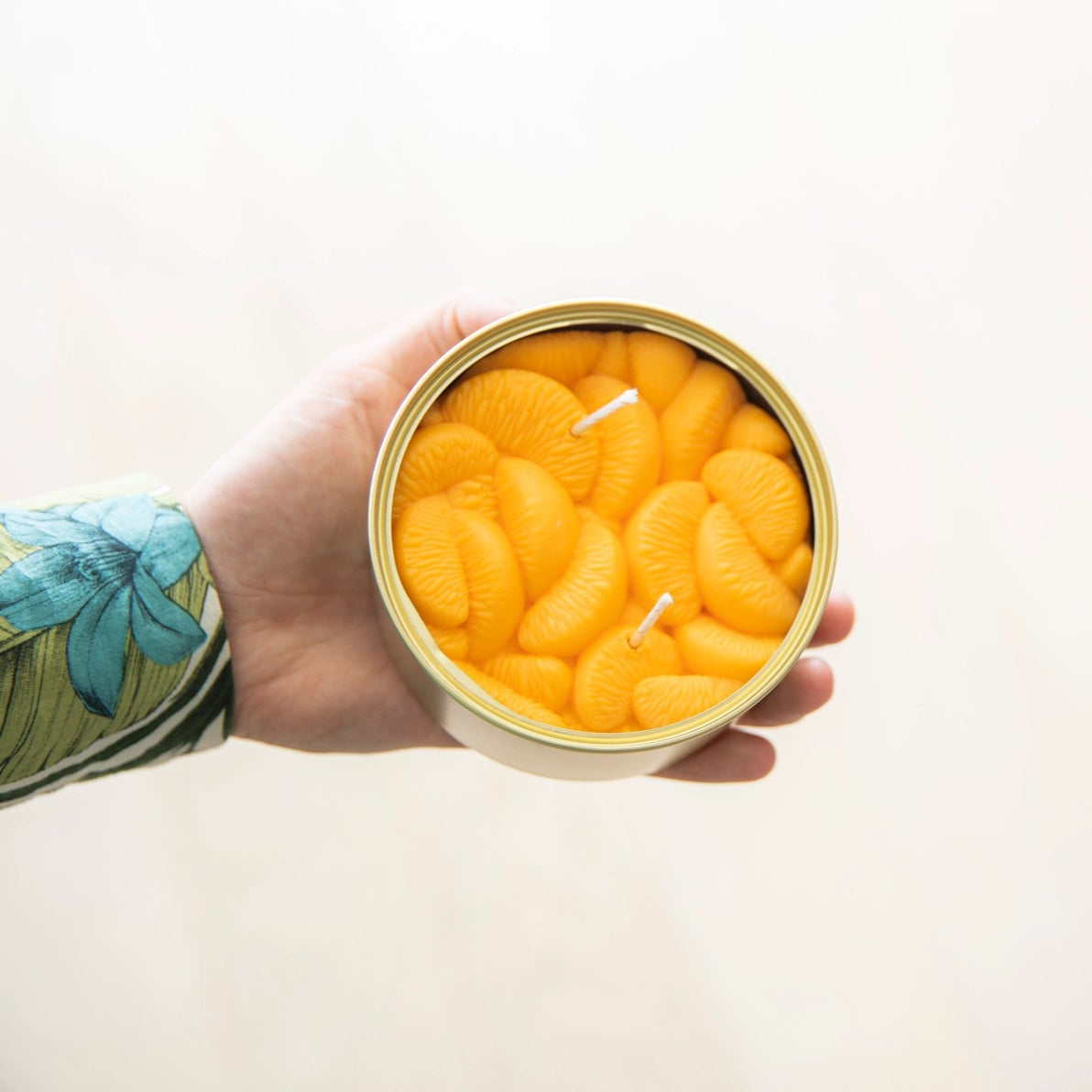 Hand holding a realistic peeled tangerine candle in an open tin, showcasing the wax segments’ lifelike texture and colour.