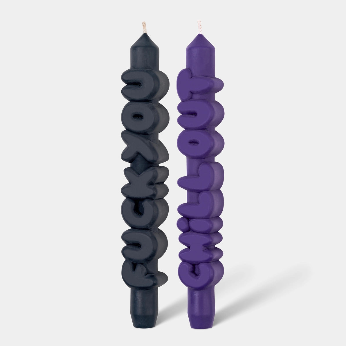 Front view of black “F*ck You” and indigo “Chill Out” taper candles with playful bubble lettering, standing upright on a light grey background.