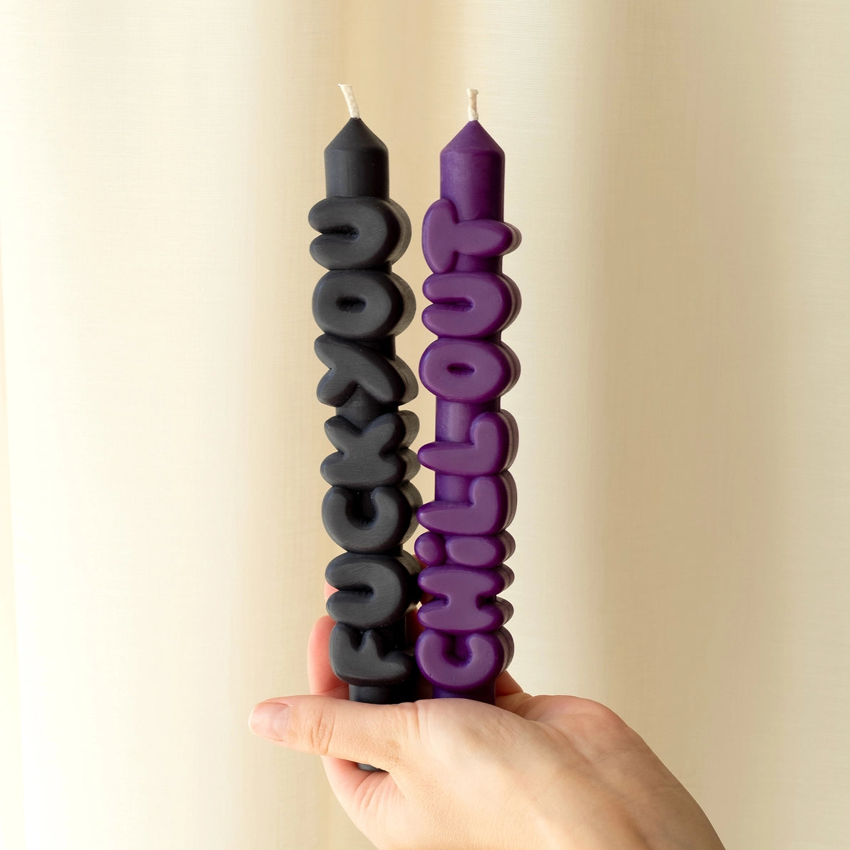 Hand holding black “F*ck You” and indigo “Chill Out” sculptural text candles in front of a cream curtain, showing off their bold shape and colour contrast.