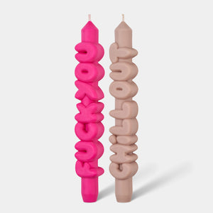 Front-facing view of pink “F*ck You” and cream “Chill Out” taper candles with bold bubble lettering, standing upright on a plain light grey background.