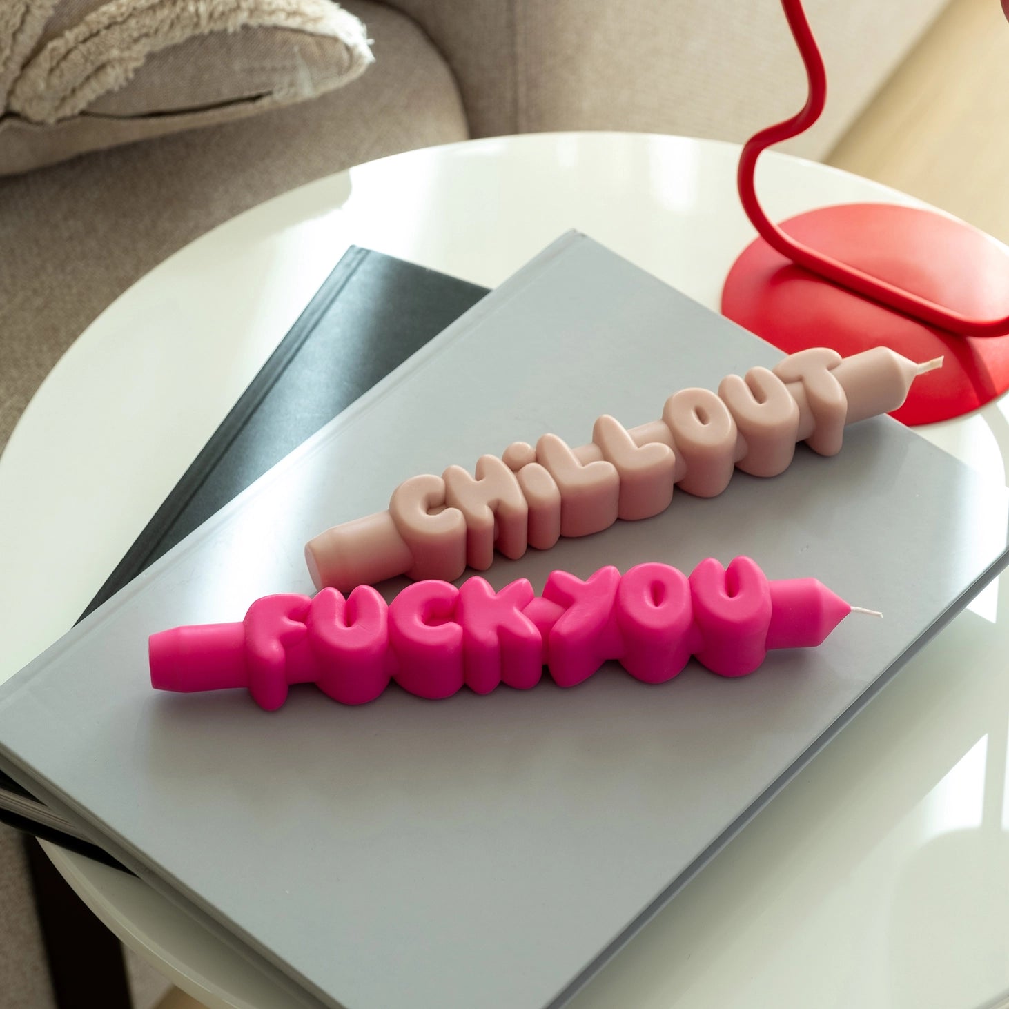 Flat lay of pink “F*ck You” and cream “Chill Out” text candles displayed on a coffee table next to a red lamp and books in a modern living room setting.