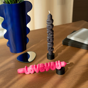 Styled flat lay of black “F*ck You” candle upright in a holder and pink “Love You” candle lying flat on a wooden table, with a wavy blue vase and mirror decor in the background.