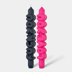 Front view of black “F*ck You” and pink “Love You” sculptural taper candles with chunky bubble text, standing upright on a light grey background.