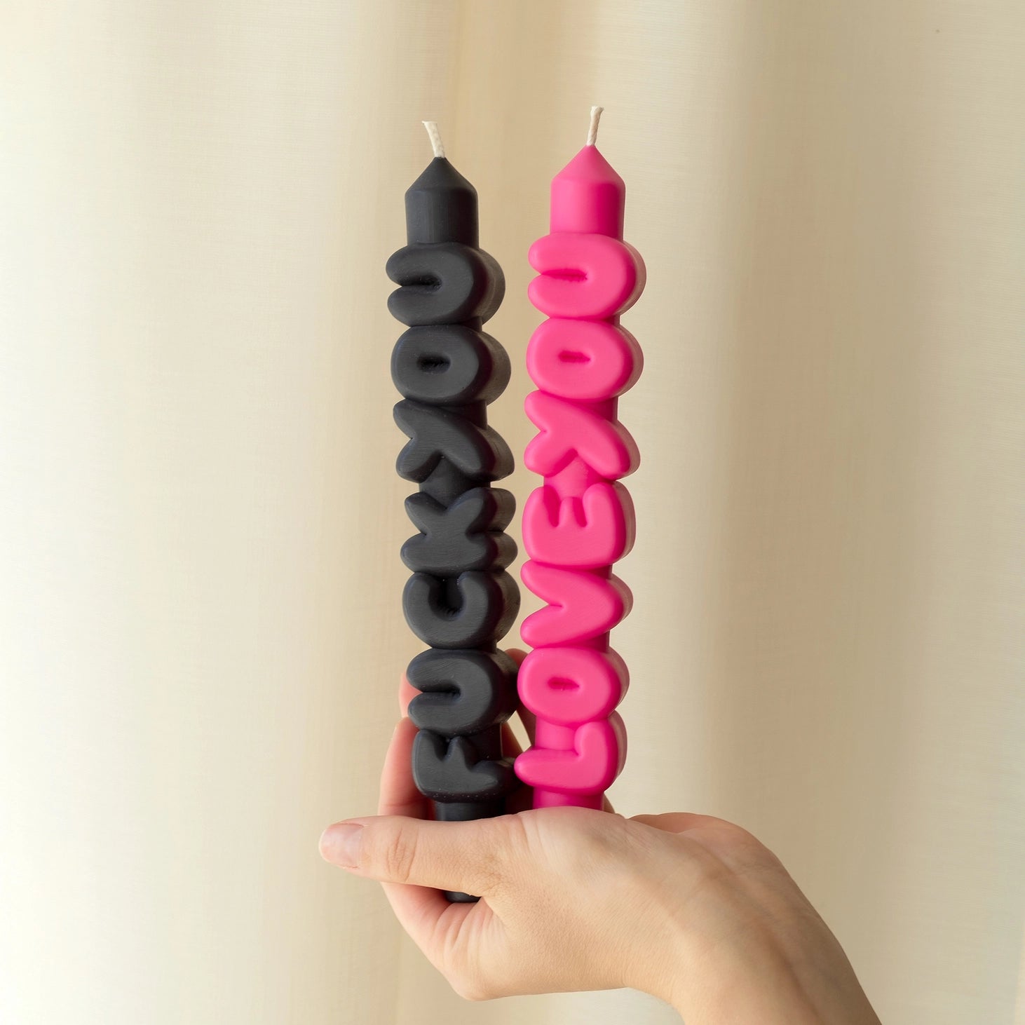 Hand holding black “F*ck You” and pink “Love You” text taper candles in front of a soft cream curtain, showcasing the bold lettering and vibrant colours.