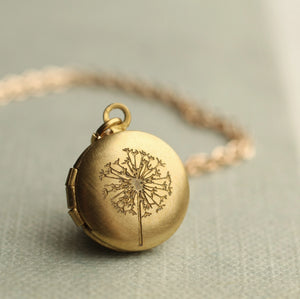 Close-up of Tiny Brass Dandelion Locket Necklace with gold-plated chain on soft fabric background.